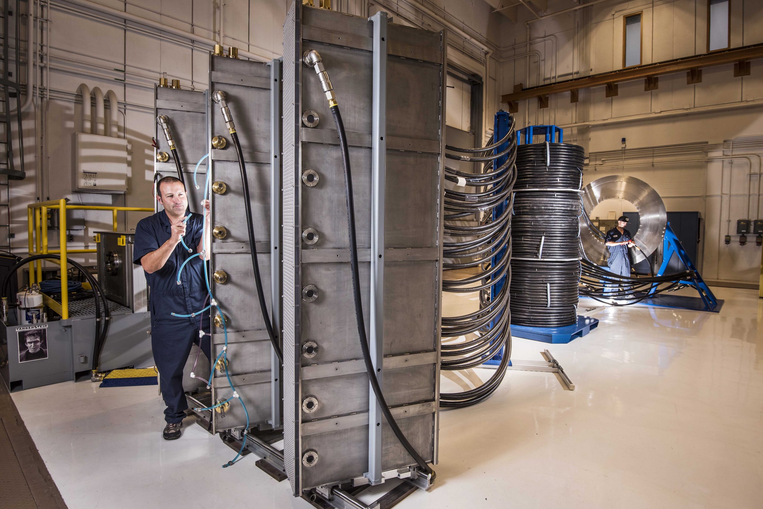 Sandia National Laboratories technician Tommy Mulville installs a gas exhaust line for a switch at Thor’s brick tower racks. In the background, beyond the intermediate support towers, technician Eric Breden makes ready an electrical cable for insertion in the central power flow assembly.