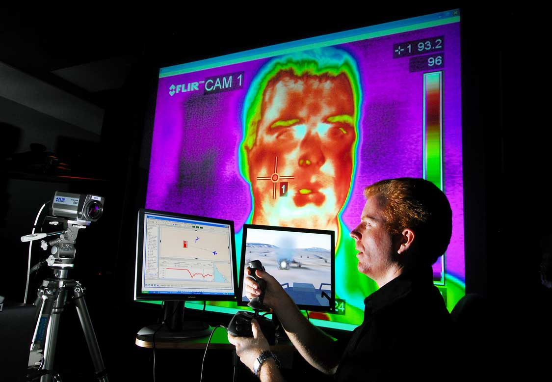 Sandia researcher Rob Abbott uses a joystick and plays the role of a student in a training exercise driving an amphibious assault vehicle simulator used by the Navy and Marines. The second monitor is an instructor/operator application called CDMTS. In the background is a thermal image of a student's face used for investigating biometrics to monitor the student in various ways including the level of engagement and focus of attention. (Photo by Randy Montoya)