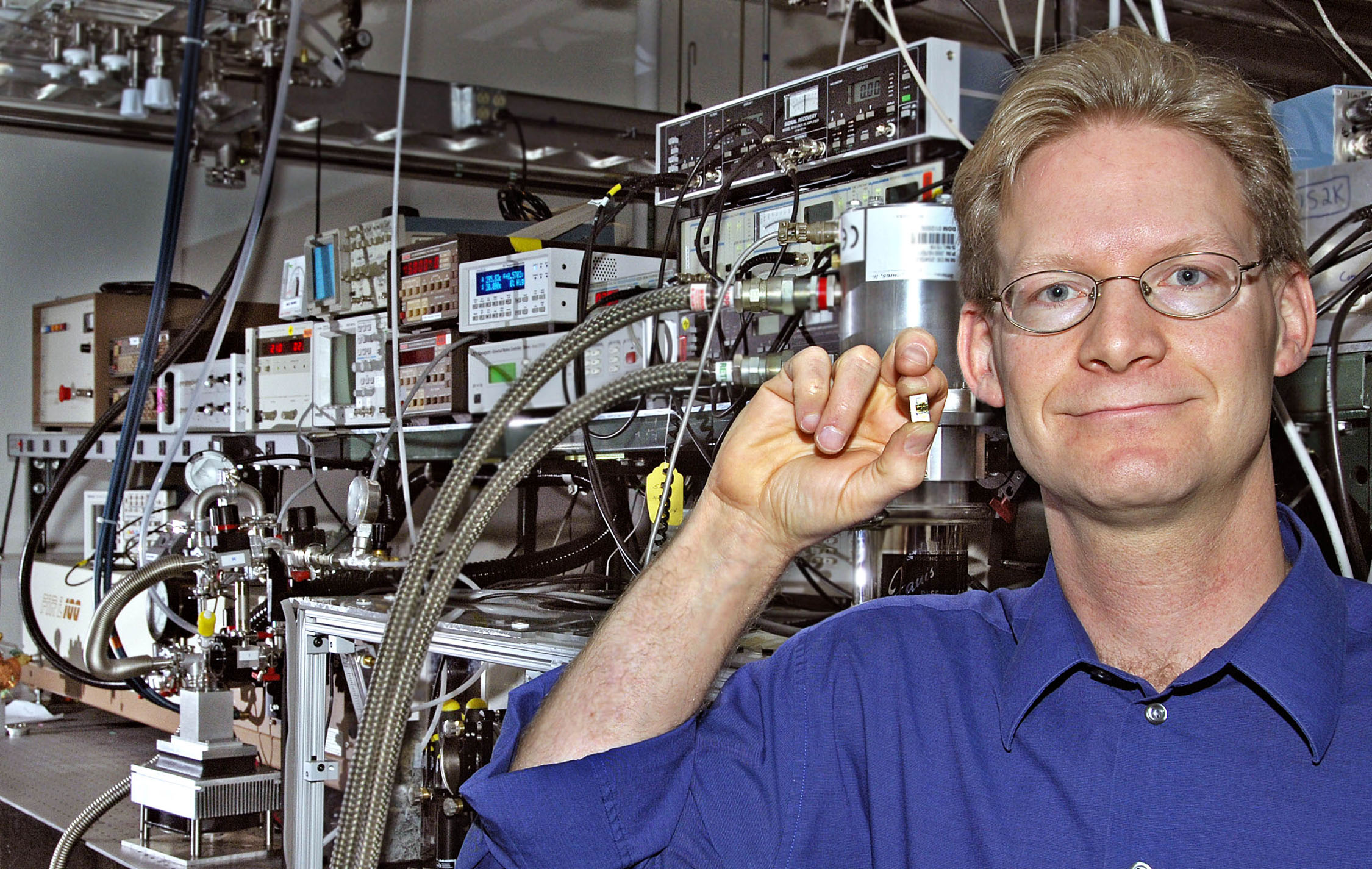 MIKE WANKE, principal investigator of the Terahertz Microelectronics Transceiver Grand Challenge, holds a miniaturized device that will eventually replace large pieces of equipment like those in the background. 