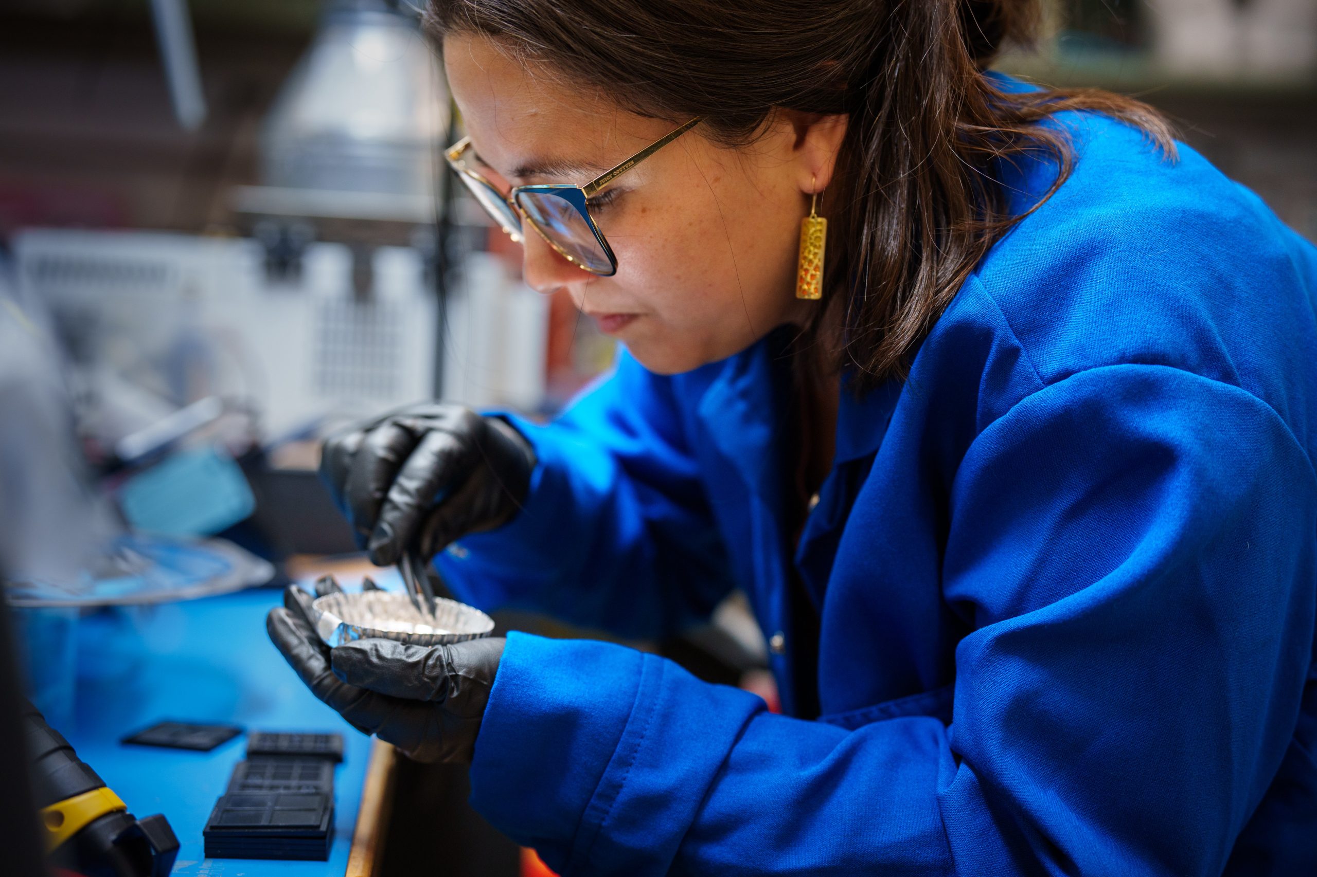 A woman in a blue lab coat grabs a tiny device with tweezers.