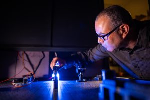 A man adjusts a science-y object with a blue light illuminating a shiny table.