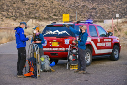 Three people with large backpacks, jackets and masks in front of a red SUV labeled Mountain Rescue