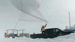 One person, in orange, stands on a pickup with a large white balloon on top, tethered to a hoist behind the truck.