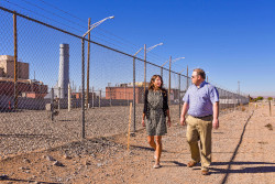 Two people walking beside a fence. A cooling stack of a nuclear facility is in the background.