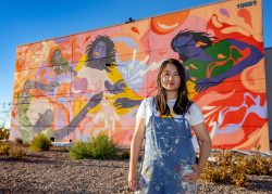 Artist Amanda Phingbodhipakkiya, in paint-splattered overalls, poses in front of her complete mural