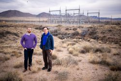 Two men stand in front of electrical grid equipment, mountains in the background.
