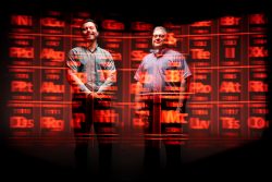 Two men stand with a red periodic table of the elements projected upon them.