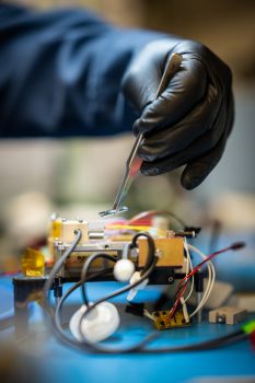 A gloved hand manipulates something with tweezers over a smallbox with wires.