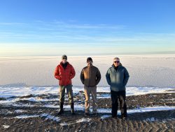 Three men in thick coats stand in front of ice.